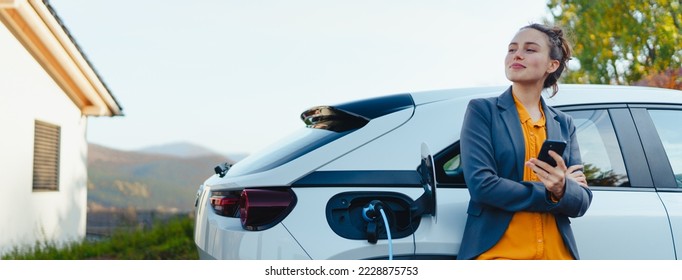 Young woman with smartphone waiting while her electric car charging in home charging station, sustainable and economic transportation concept. - Shutterstock ID 2228875753