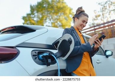 Joven con smartphone esperando mientras cargaba su auto eléctrico en la estación de carga de la casa, concepto de transporte sustentable y económico.