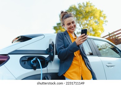 Young woman with smartphone waiting while her electric car charging in home charging station, sustainable and economic transportation concept. - Powered by Shutterstock