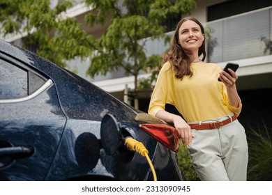 Young woman with smartphone waiting while her electric car charging in home charging station, sustainable and economic transportation concept. - Powered by Shutterstock