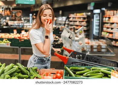 Young woman smelling a tomato while grocery shopping in a supermarket. She is pushing a shopping cart and choosing fresh produce from the shelves. Shopping concept - Powered by Shutterstock