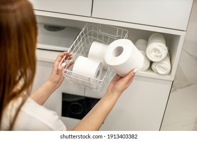Young woman with a smile is organizing and placing mesh basket with stack of toilet paper rolls in bathroom cupboard near rolled up hand towels. Happy housewife is tidying up toiletries in bathroom.