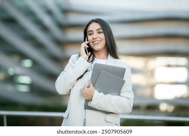 A young woman is smiling and engaged in a phone conversation while holding a laptop and notebook outdoors in a modern urban setting. - Powered by Shutterstock