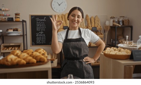 Young woman smiling inside a bakery shop wearing an apron and making an okay gesture in front of bread and pastries displays. - Powered by Shutterstock