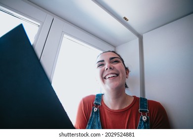 Young Woman Smiling And Laughing While Reading A Book In The Window, Bright Day, Relax And Mental Health Concepts