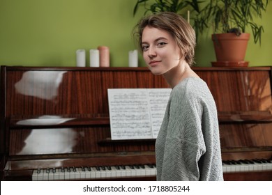 Young woman smiling looking at the camera sitting near acoustic piano with music sheet. Home islolation. Feeling comfortable at home. Flowers in flowerpots on a piano. Lifestyle at quarantine.