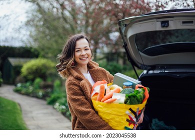 A Young Woman Smiling, Looking At The Camera And Holding A Reusable Carrier Bag Filled With Groceries. She Is Unloading The Car After Food Shopping.