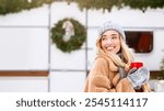 A young woman, smiling radiantly, holds a warm drink while dressed in cozy winter attire. She stands near holiday decorations, capturing the essence of a joyful winter day.