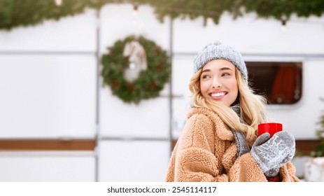 A young woman, smiling radiantly, holds a warm drink while dressed in cozy winter attire. She stands near holiday decorations, capturing the essence of a joyful winter day. - Powered by Shutterstock