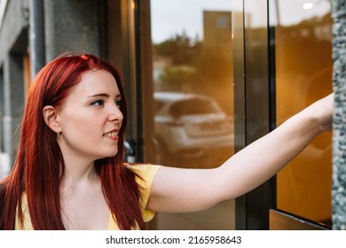 Young Woman Smiling, Ringing The Doorbell Of A Friend's House In A Block Of Buildings In A Big City. Girl Stops By To Pick Up Her Friend. Concept Of Friendship.