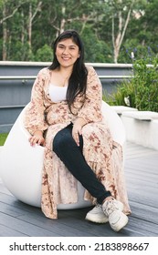 Young Woman Smiling, Sitting On A Chair In The Mall, In A Green Area. Portrait Of A Colombian Girl Looking At The Camera. Woman Dressed In Jeans, Blouse And Tennis Shoes.