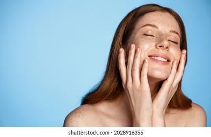 Young Woman Smiling, Touching Glowing Hydrated Skin With Pleasure, Hands On Natural, Healthy Face, Close Eyes And Big Smile, Stands Against Blue Background