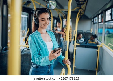 Young woman smiling while standing by herself on a bus and listening to music on a smartphone. Happy female passenger using headphones and a mobile phone in public transportation. Copy space. - Powered by Shutterstock