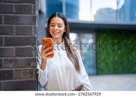 Young woman smiling while using smartphone outdoors, confident and connected in an urban setting, checking messages, browsing apps and engaging with social media during daytime walk