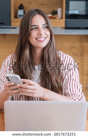Young woman smiling while using a smartphone and laptop in a cozy home office kitchen, remote work and casual striped shirt conveying relaxed productivity and modern lifestyle