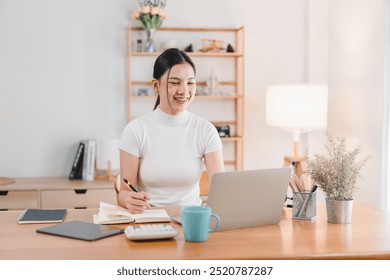 A young woman is smiling while working on her laptop at cozy home office. She is taking notes and appears focused and happy in her bright, well organized workspace. - Powered by Shutterstock