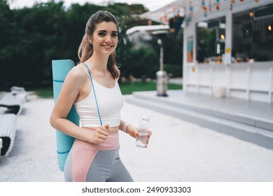 Young woman smiling with a yoga mat and a bottle of water, ready for a yoga session in a park. She stands near an outdoor café with colorful decorations, embodying a healthy and active lifestyle - Powered by Shutterstock