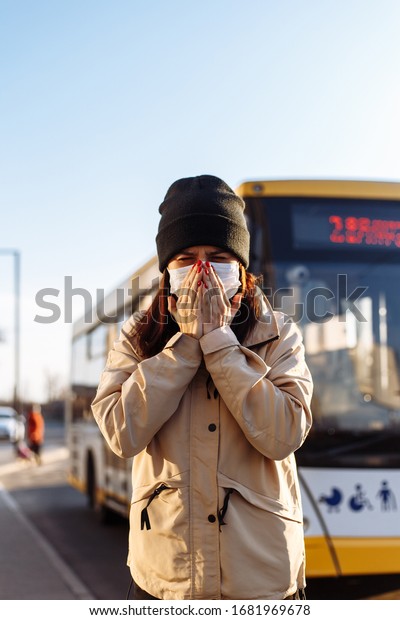 Young woman sneezes on a bus station and covers\
her face with hands. Girl wears a white medical mask standing near\
a bus at a public transport station. Coronavirus illness concept.\
Pandemia, epidemia.
