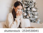 Young woman sneezing into a tissue, sitting by a festive Christmas tree. The cozy scene conveys seasonal allergies or cold symptoms during the winter holiday season, with warm, comforting ambiance.