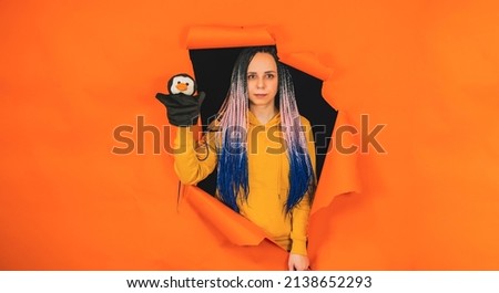 Young woman with soft puppet toy on hand looking out of hole of orange background. Pretty female with puppet penguin. Concept of puppet show