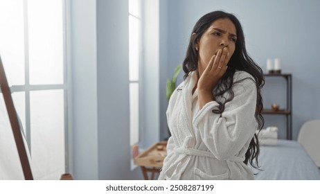 Young woman in a spa wearing a white robe, looking tired in an indoor wellness center with minimalist decor, near large windows and a massage table. - Powered by Shutterstock