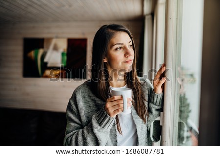 Young woman spending free time home.Self care,staying home.Enjoying view,gazing through to the window.Quarantined person indoors.Serene mornings.Avoiding social contact.