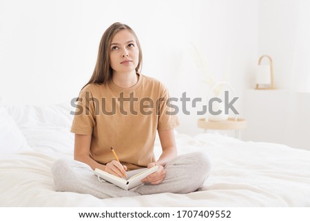 Young woman spending free time in bedroom, sitting on bed cover, thinking of plans for week and writing them in notebook that is on her knees