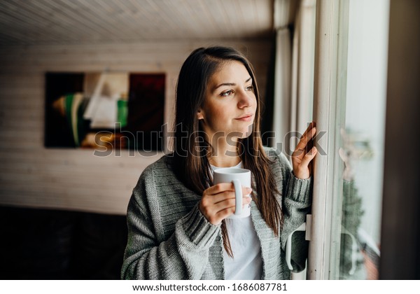 Young woman spending\
free time home.Self care,staying home.Enjoying view,gazing through\
to the window.Quarantined person indoors.Serene mornings.Avoiding\
social contact.