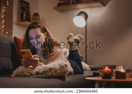 A young woman spends a cozy winter evening at home lying on the sofa looking at her smartphone. Winter holidays, Christmas and online surfing 