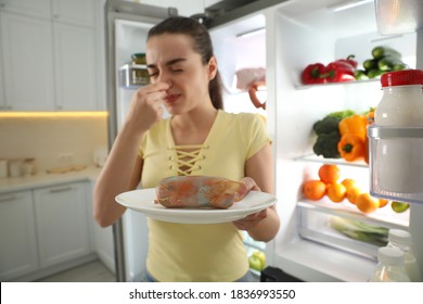 Young Woman With Spoiled Sausage Near Open Refrigerator In Kitchen