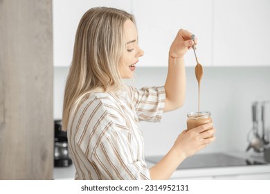 Young woman with spoon and jar of nut butter in kitchen - Shutterstock ID 2314952131
