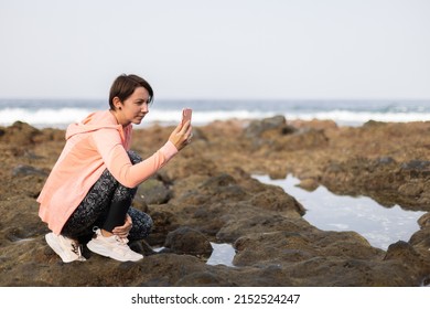 Young Woman In Sport Clothes Walking Near Ocean And Making Photos.