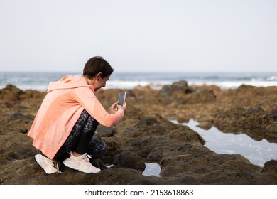 Young Woman In Sport Clothes Walking Near Ocean And Making Photos.