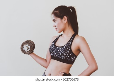 Young woman at the sport gym doing arms exercises on a machine on the background of the partition. She wears dark sportswear.
