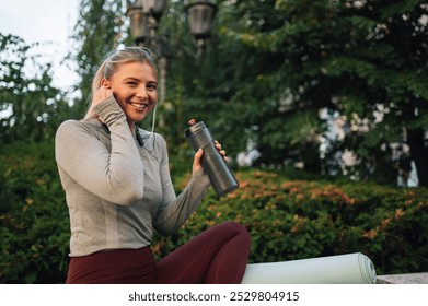 Young woman in sportswear adjusts her earphones and sips water during a park workout, looking happy and relaxed in green surroundings - Powered by Shutterstock