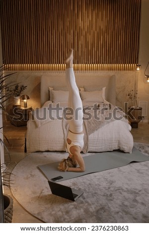 Young woman in sportswear doing yoga in supported headstand pose in bedroom with laptop on floor