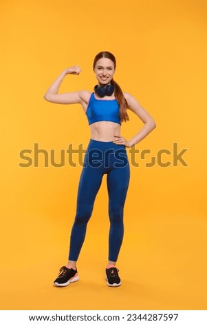 Young woman in sportswear and headphones showing muscles on yellow background