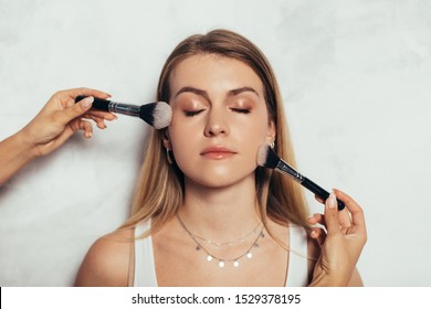 Young Woman Stand With Eyes Closed. Somebody's Hands Put Make Up On Her Face With Brush. Beauty Routine. Isolated Over White Background