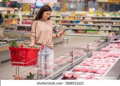 Young Woman Standing In Aisle With Shopping Cart Choosing Fresh Raw Meat In Modern Supermarket