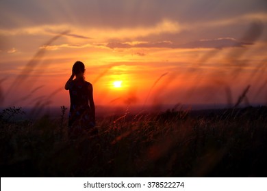 Young Woman Standing Alone In A Field During Sunset. Silhouette Of A Young Girl Outdoor.