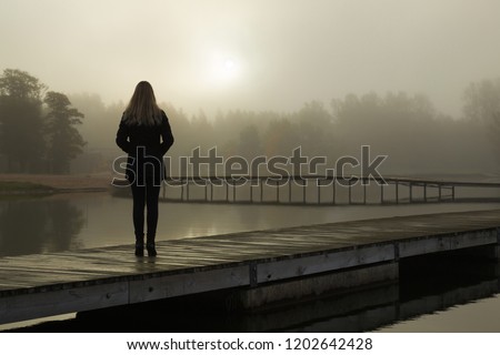 Young woman standing alone on lake footbridge and staring at sunrise in gray, cloudy sky. Mist over water. Foggy air. Early chilly morning. Dark, scary moment and gloomy atmosphere. Back view.