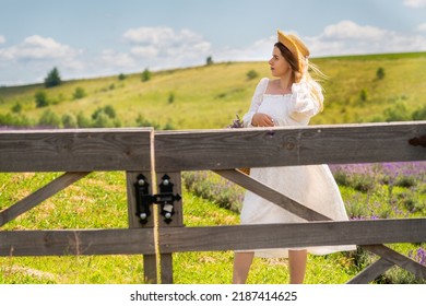 Young Woman Standing Behind A Closed Rustic Farm Gate In Fresh White Cotton Dress And Straw Sunhat Looking Off To The Side