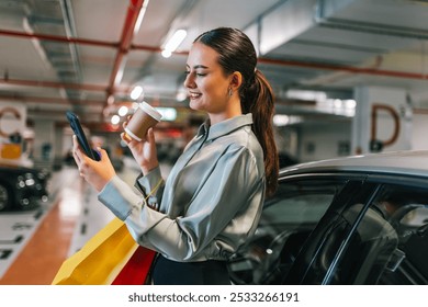 Young woman standing by her car with shopping bags using her phone and drinking coffee. - Powered by Shutterstock