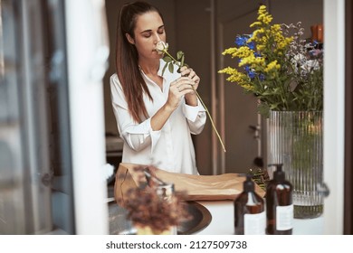 Young woman standing at a counter in her kitchen at home smelling freshly cut flowers while putting them into a glass vase