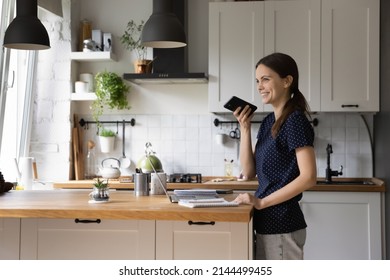 Young woman standing in domestic kitchen holding smart phone leaves voice message, share voicemail to friend, having pleasant conversation use speakerphone, easy convenient modern tech usage concept