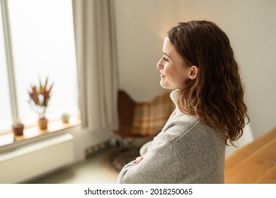 Young Woman Standing With Folded Arms Gazing Out Of A Window With A Smile Indoors At Home In A Close Up Side View With Copyspace