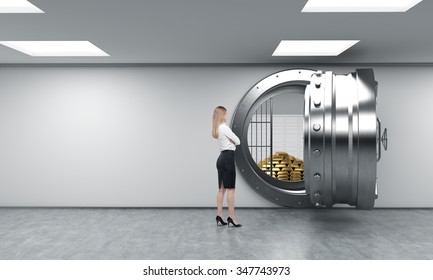 Young Woman Standing In Front Of A Big Unlocked Round Metal Safe In A Bank Depository With A Pyramid Of Gold Bars And Lock-boxes Inside,  A Concept Of Security And Client Service