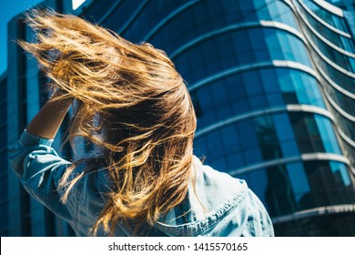 Young Woman Standing In Front Of Blue Building Of Business Center, Her Hair Fluttering In Wind, Rear View.