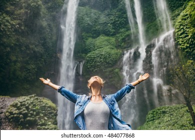 Young Woman Standing In Front Of Waterfall With Her Hands Outstretched. Caucasian Female Tourist In Forest With Her Arms Wide Open.