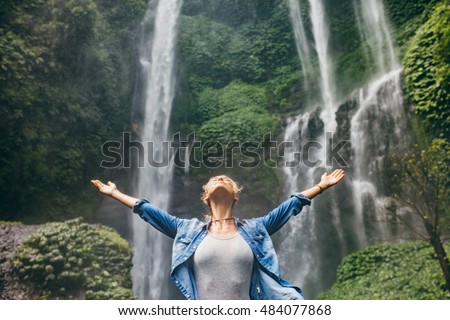 Young woman standing in front of waterfall with her hands outstretched. Caucasian female tourist in forest with her arms wide open.
